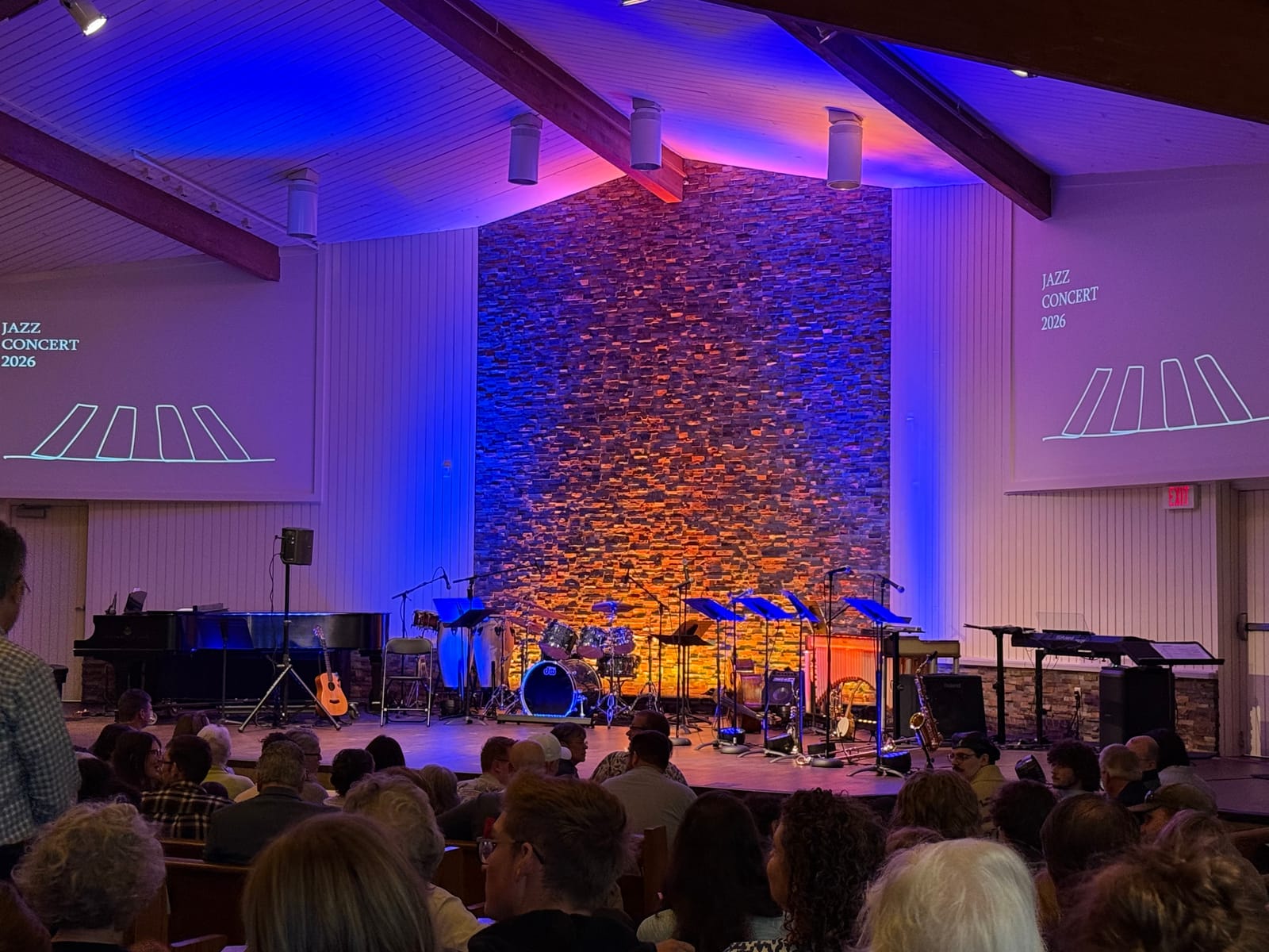 Audience seated before the lit stone-wall stage at University Reformed Church’s 2026 Jazz Concert, with instruments arranged on stage and “Jazz Concert 2026” projected on the side walls.